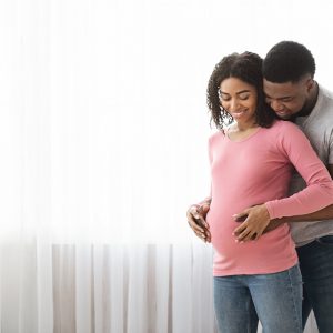 Loving pregnant black couple cuddling at home, standing next to window, copy space