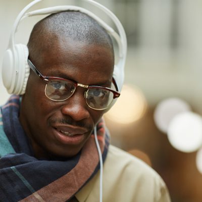 Head and shoulders portrait of trendy African man wearing headphones looking at camera while posing in beautiful building outdoors, copy space